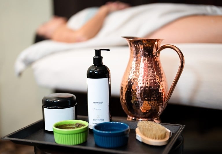 A black tray holds a pump bottle, jar, two small bowls, a brush, and a hammered copper pitcher, set before a person on a massage bed.
Text: HERBAL BODY OIL Rejuvenating Action For all skin types With Almond & Apricot Kernel Oil for all skin types FOR PROFESSIONAL USE
