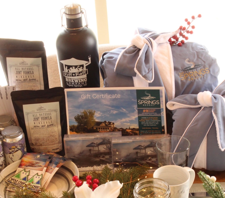 A collection of promotional items and gifts, including a husky toy, bathrobes, a beer growler, and mineral soaks, are displayed on a wooden surface.