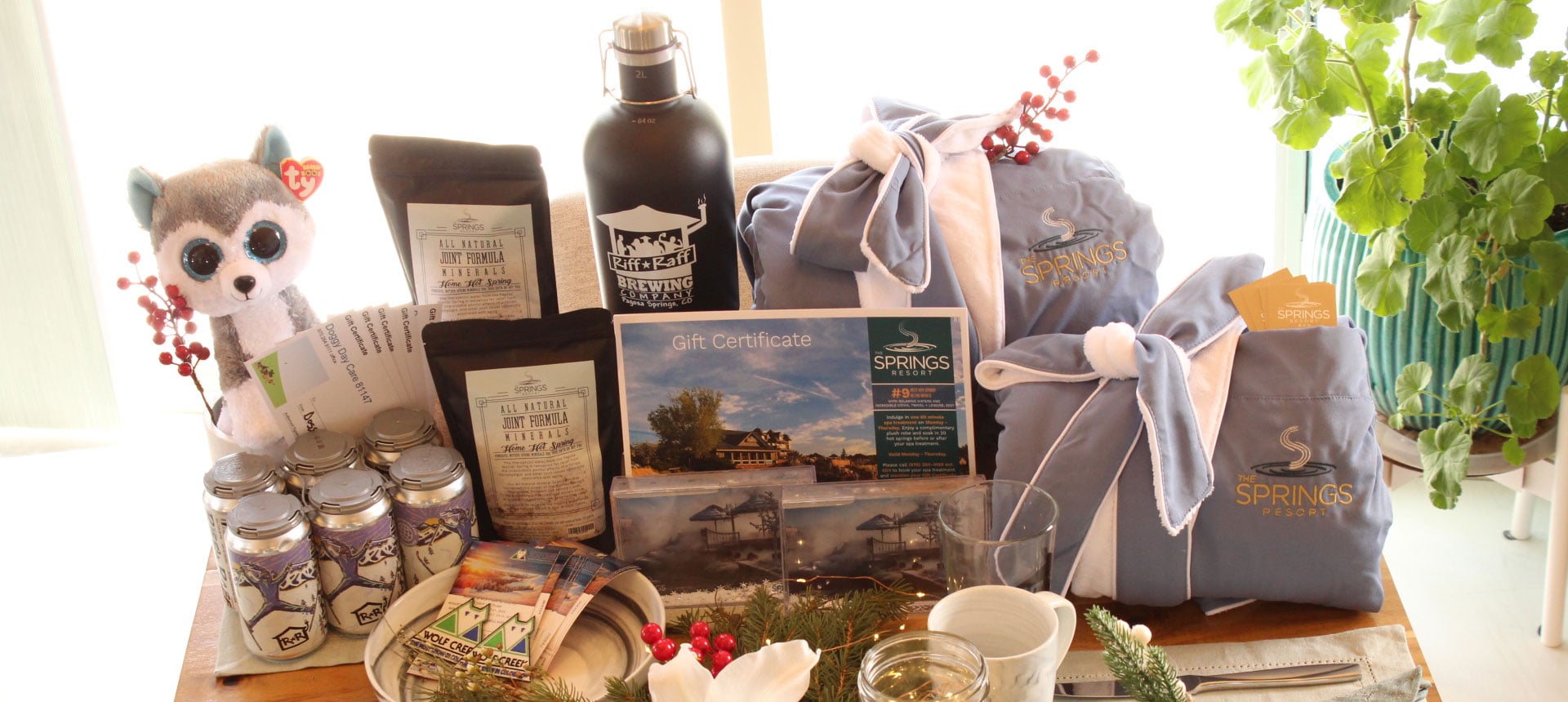A collection of promotional items and gifts, including a husky toy, bathrobes, a beer growler, and mineral soaks, are displayed on a wooden surface.