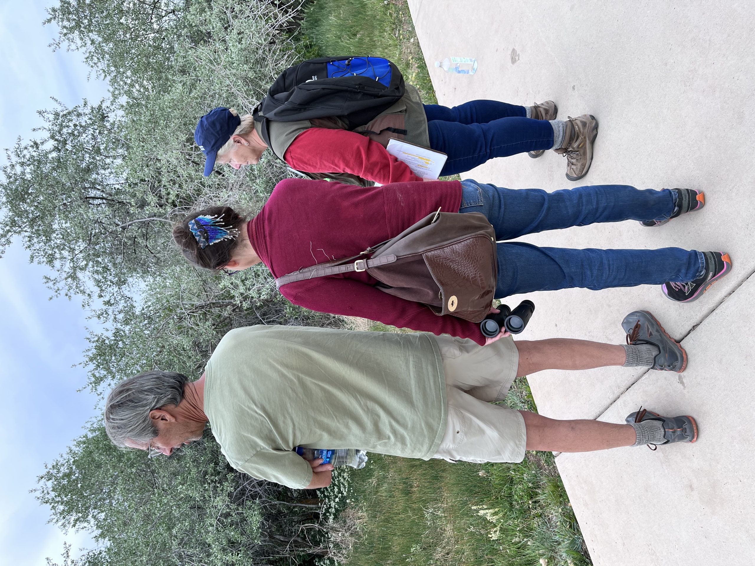 Three people stand on a concrete path beside green bushes. A man holds an AQUAFINA bottle; a woman holds binoculars and wears NRS shoes; another woman wears a NATIONAL backpack. Another AQUAFINA bottle is on the ground.