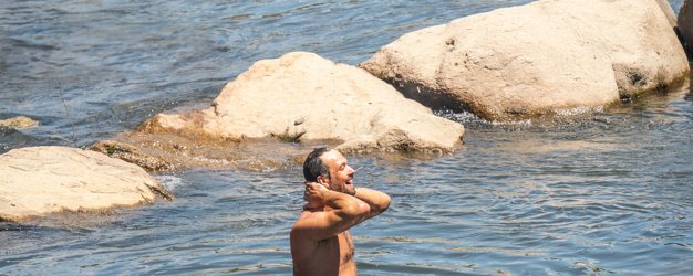 A shirtless man stands chest-deep in rippling water, hands behind his head, smiling upwards. Several large rocks surround him in the clear river.
