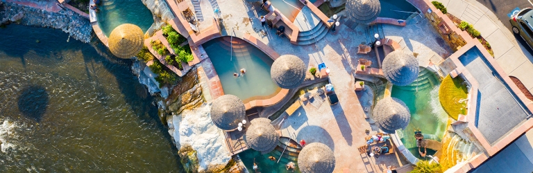Terraced hot spring pools with people bathing and relaxing under straw umbrellas. The complex is integrated into a rocky riverbank bordering a dark river, with surrounding pathways.