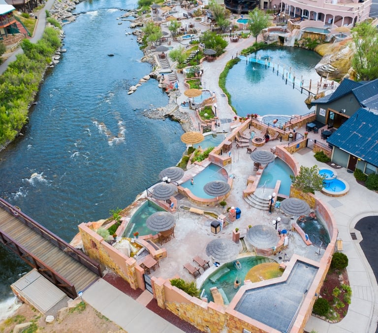 A sprawling hot springs complex features numerous pools with people relaxing under straw umbrellas. It's built along a wide river with a pedestrian bridge, bordered by buildings and lush greenery.