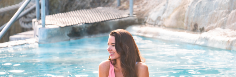 A woman smiles, leaning on a rustic pool edge, submerged chest-deep in steaming blue water, with metal steps and rocky terrain behind her.