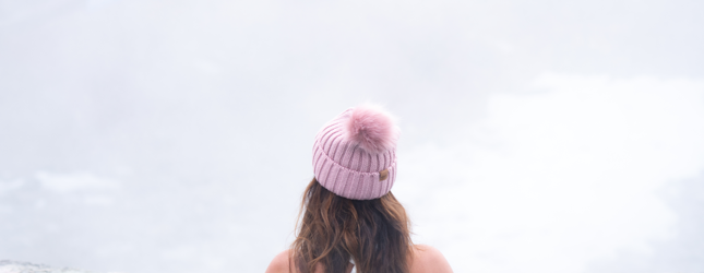 A woman in a pink pom-pom hat and white swimsuit stands chest-deep in a blue pool, holding the edge, looking out at a bright, misty background.