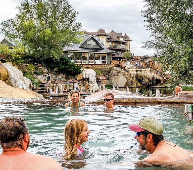 A diverse group relaxes in a natural hot spring pool surrounded by rock formations and multi-story buildings, one featuring a 