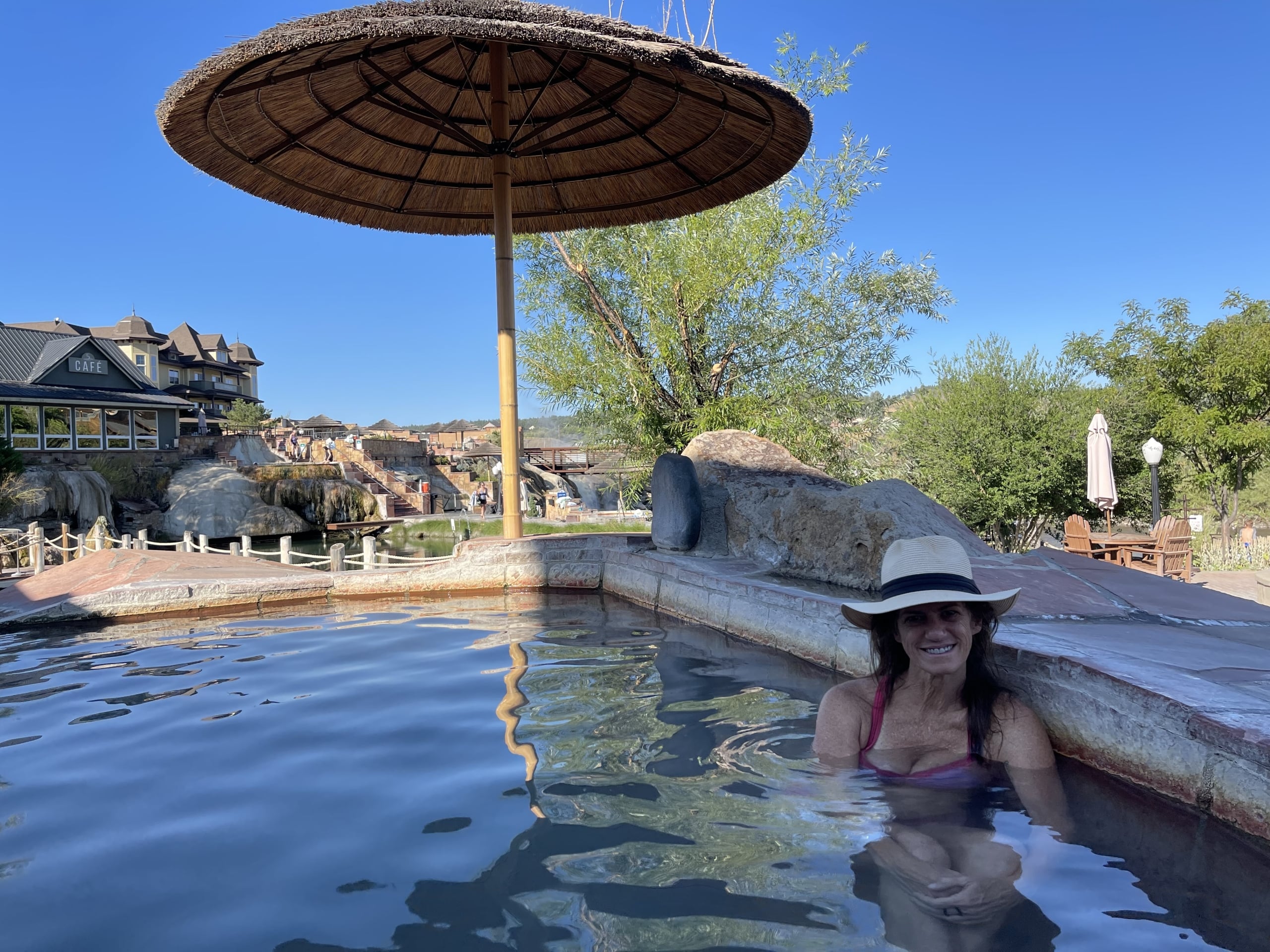 A woman smiles while soaking in a hot spring pool under a large straw umbrella. A building labeled 