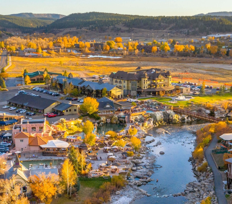 Resort complex and small town with buildings, parking lots, and thermal pools bordering a river, surrounded by autumn foliage and forested mountains. Text: VAIDA, THE Springs.