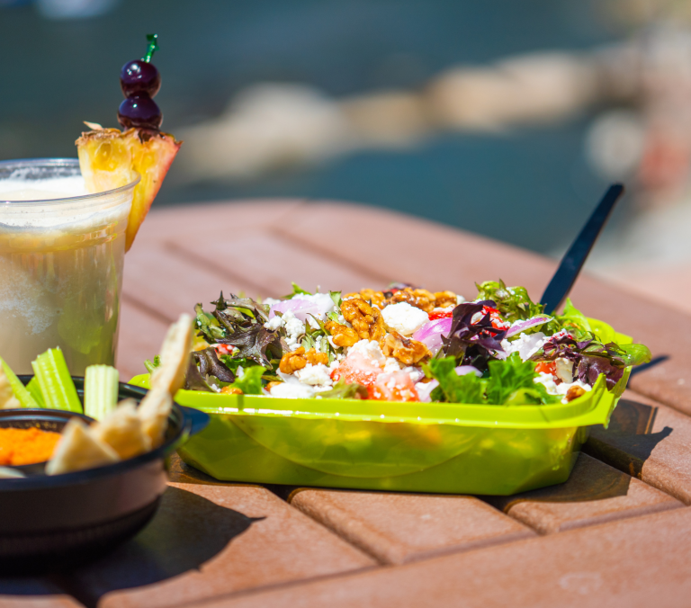 A vibrant salad, a blended drink garnished with pineapple and cherries, and pita with dip sit on a slatted brown outdoor table under sunlight with a blurred water background.