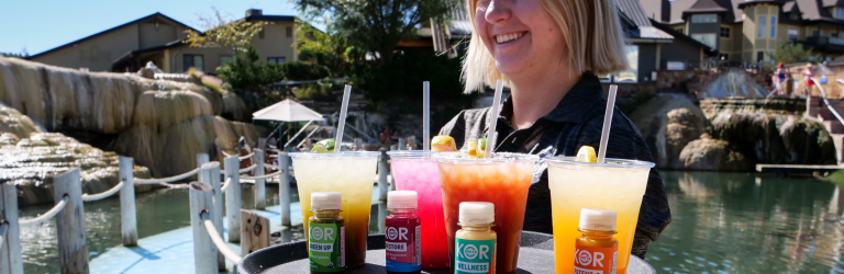 A smiling woman carries a tray with four colorful drinks and four small KOR SHOTS supplement bottles on a walkway beside water, with waterfalls and buildings in the background.
