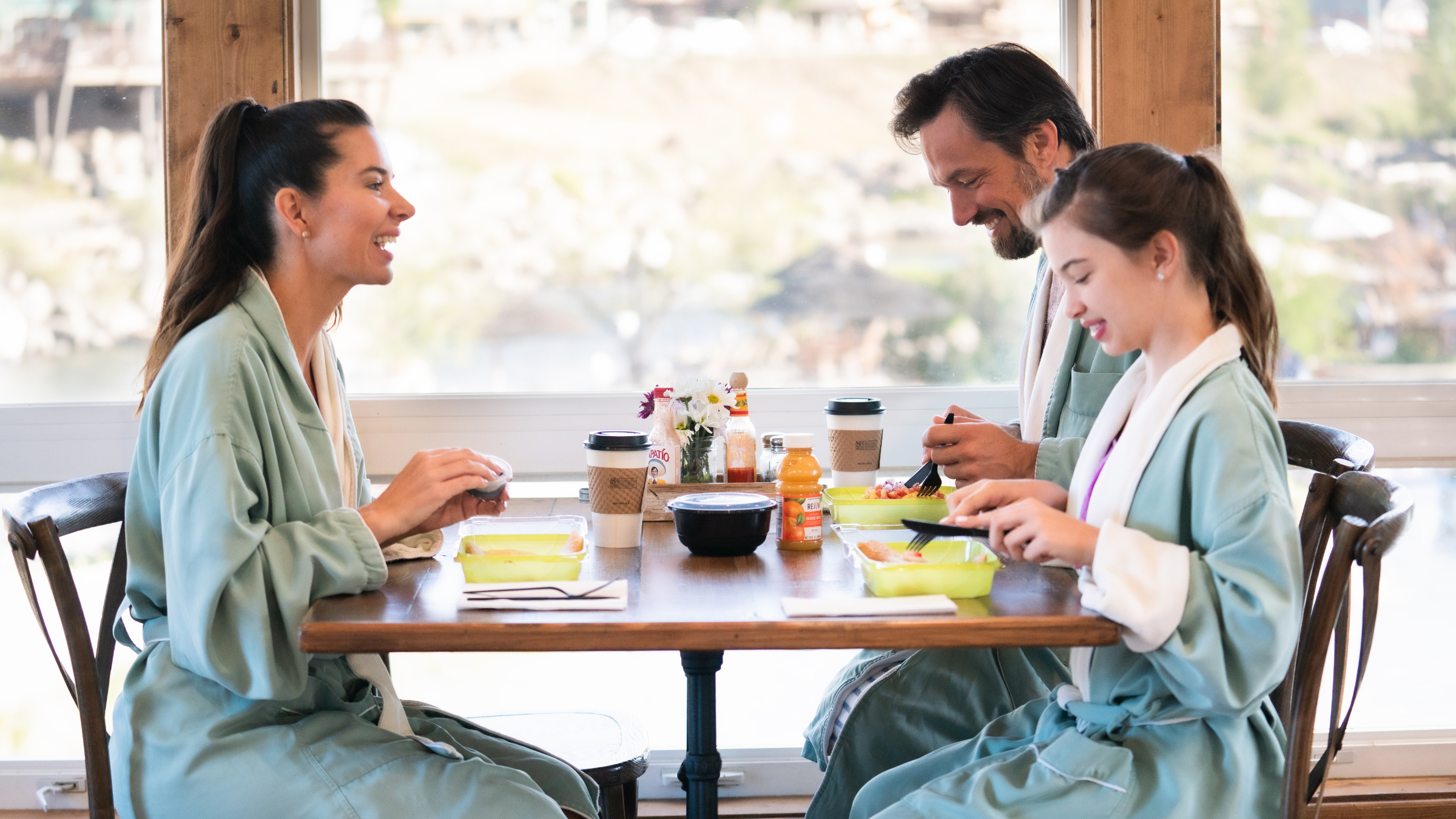 Three people in teal robes eat from takeout containers at a wooden table, smiling in front of a large window with an outdoor view. Text: RECYCLABLE, PATIO, ORANGE.