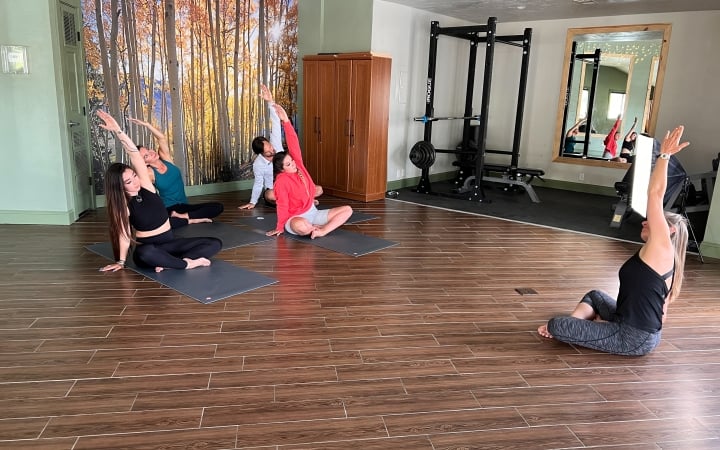Four people are seated on yoga mats, stretching with arms extended in a studio with wood-tiled floors, a birch tree mural, and 