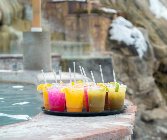 A black tray holds colorful drinks with straws and fruit, resting on a stone wall beside a steaming pool, backed by a snowy rock formation.