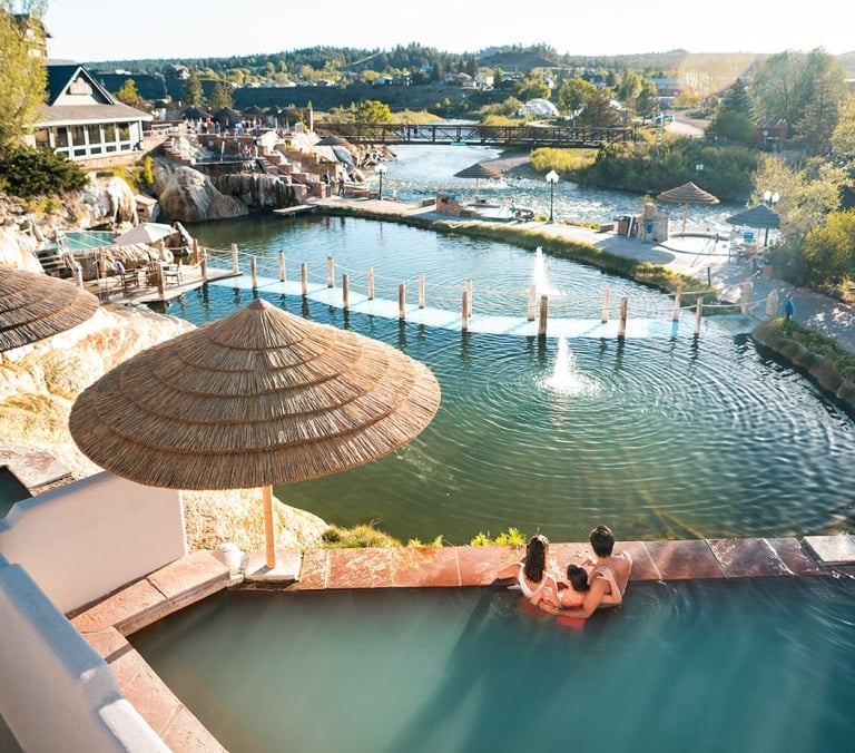 A family of four relaxes in a terraced outdoor hot spring pool, overlooking a large complex of connected pools and a river with a bridge, all under a sunny sky.
