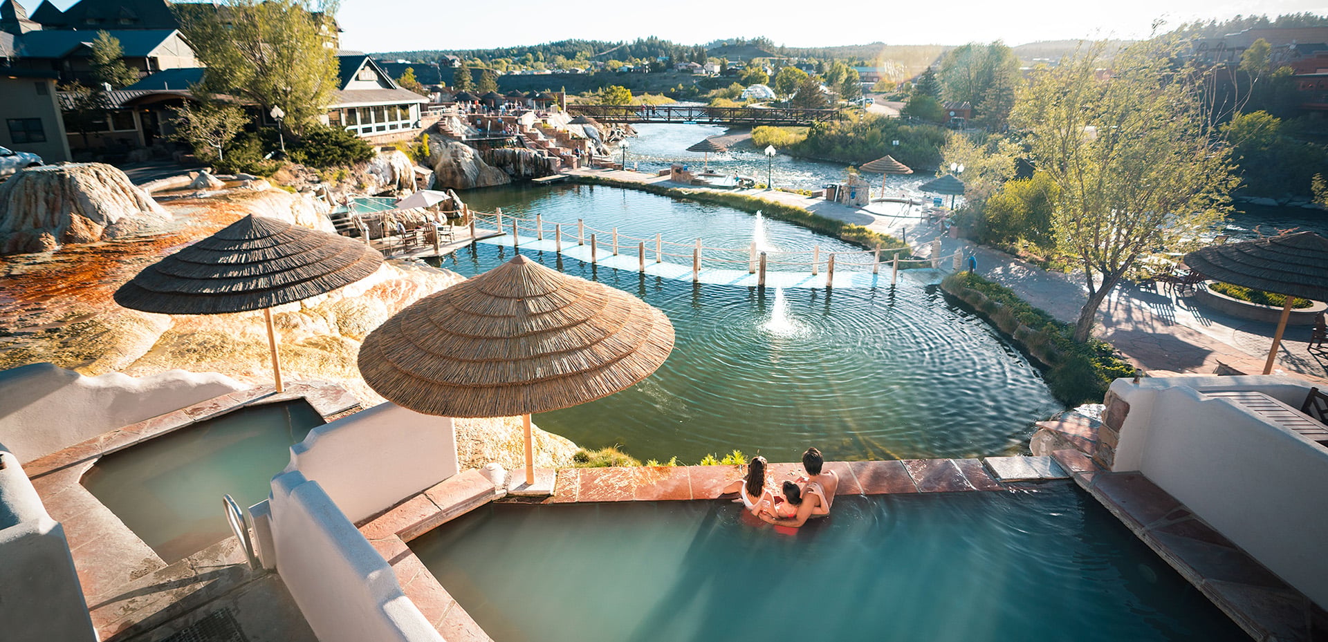 A family of four relaxes in a terraced outdoor hot spring pool, overlooking a large complex of connected pools and a river with a bridge, all under a sunny sky.