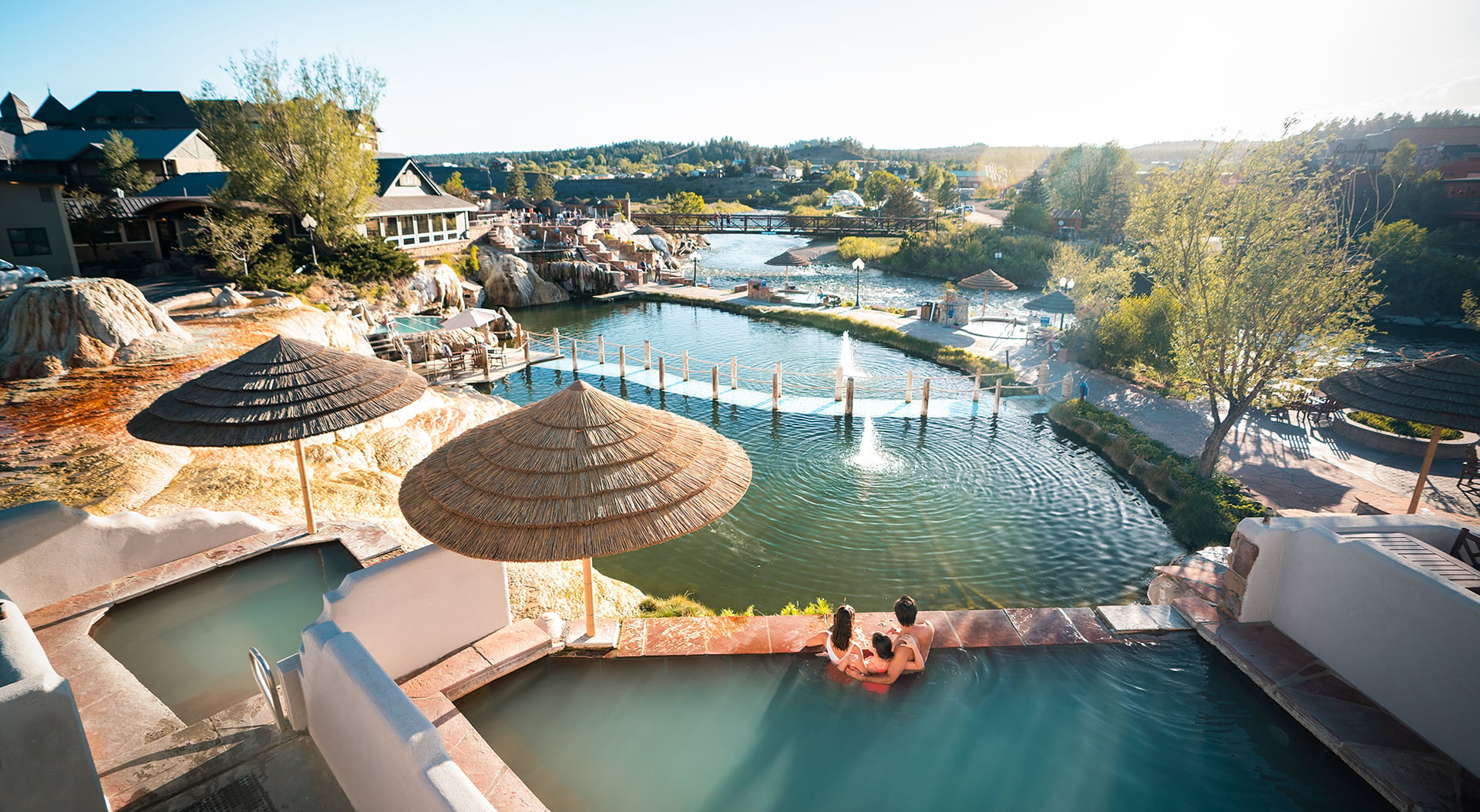 A family relaxes in a hot spring pool overlooking a complex of natural pools, a river with a bridge, and buildings under a bright sunny sky.