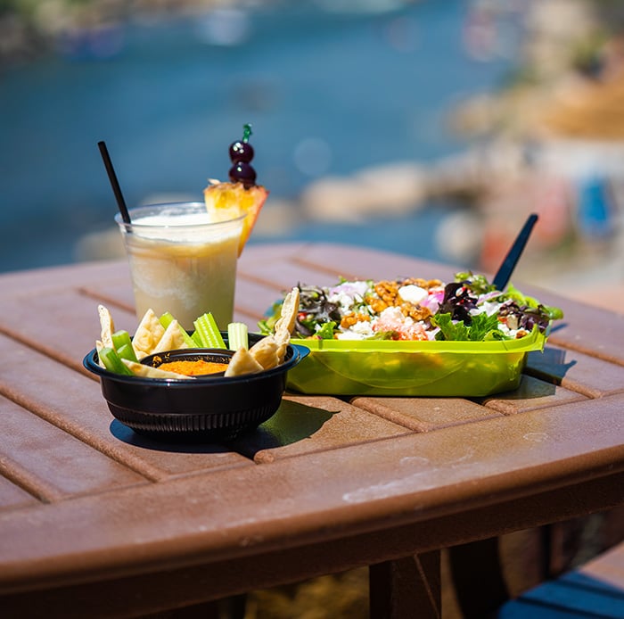 A salad, dip with celery and crisps, and a blended drink sit on a brown outdoor table, overlooking a blurred blue river under sunlight.