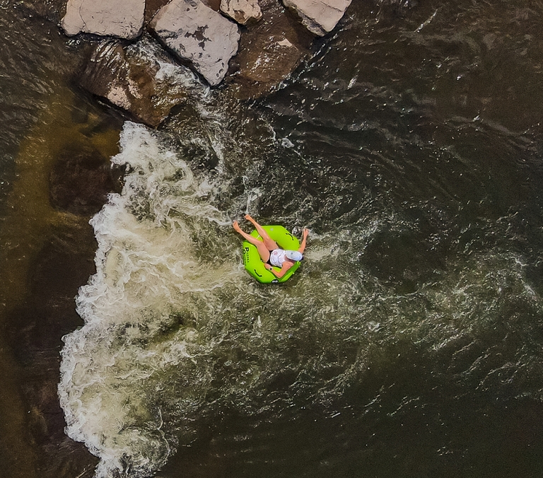 A person floats in a neon green inner tube, legs outstretched, down a dark river with white rapids and rocks nearby. Text: PACIFIC HYDRO PRO SERIES