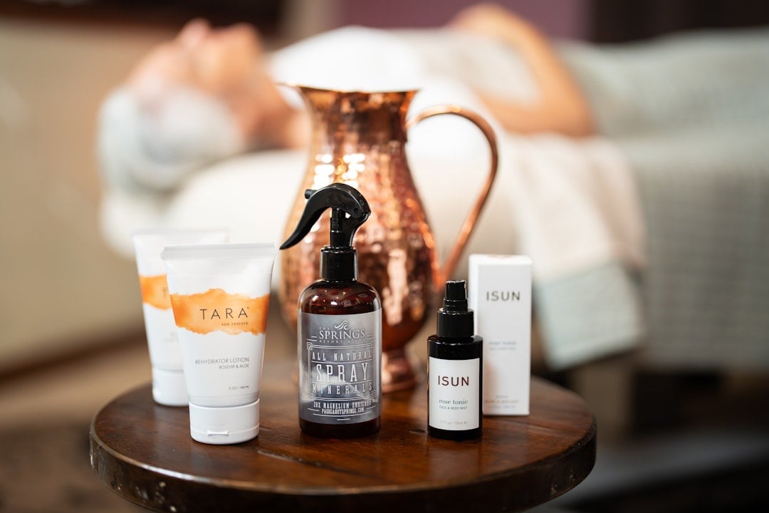 Various spa products, including lotions and sprays, stand on a wooden table before a blurred person on a massage table and a copper pitcher.
Text: TARA spa restore REHYDRATOR LOTION ROSEHIP & ALOE 3 OZ / 90 ML. THE SPRINGS RESORT ALL NATURAL SPRAY MINERALS 20X MAGNESIUM ENRICHED PAGOSASPRINGS.COM. ISUN rose tonic FACE & BODY MIST 1.7 FL OZ / 50 ML. ISUN rose tonic FACE & BODY MIST 1.7 FL OZ / 50 ML.
