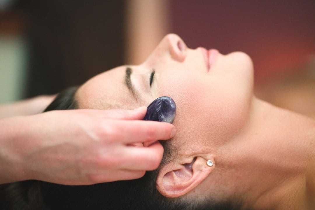 A person's serene profile, eyes closed, has a hand gently pressing a smooth, dark purple stone against their right cheek, suggesting a facial treatment.