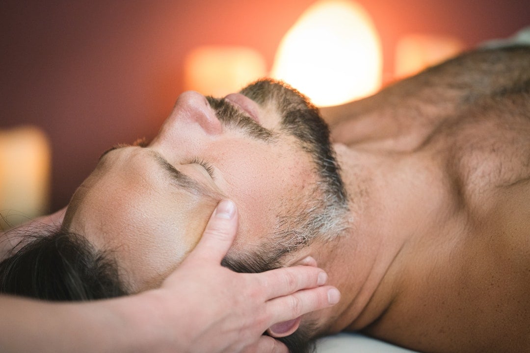 A bearded man, eyes closed, receives a head massage from hands. He lies on his back in a dimly lit room with warm, blurred lights in the background.