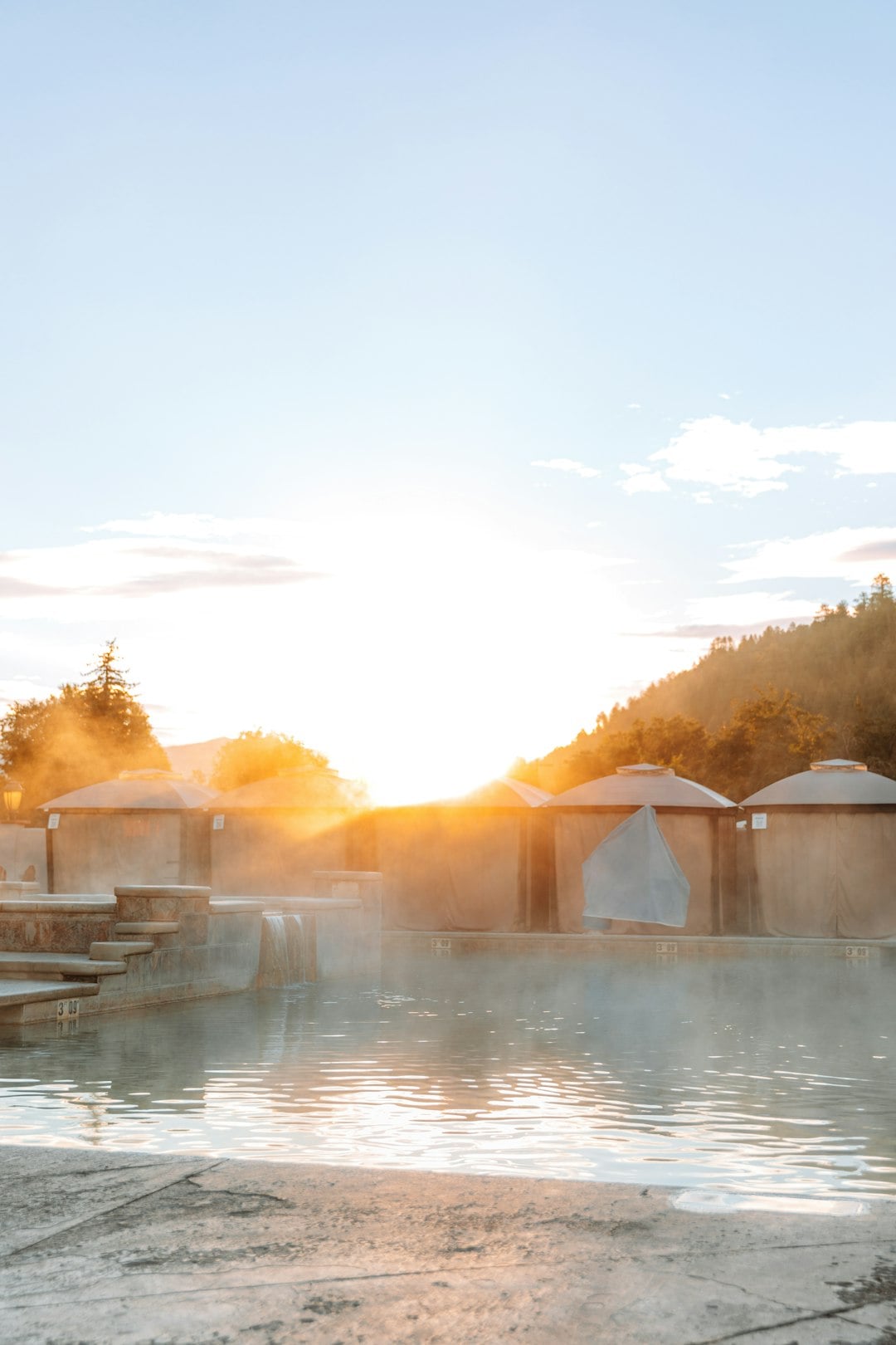 Steaming hot spring pools cascade between tiered sections, bathed in bright golden sunlight. Private cabanas and a forested mountain stand behind. Text: 3 09, 3 07, 3 09.