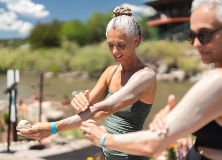 Two women apply a thick, light-colored substance to their arms, smiling. They are outdoors by a body of water, surrounded by green foliage and a building, under a sunny sky.