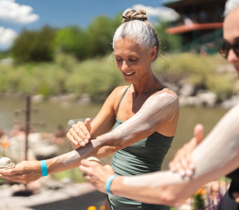 Two women apply a thick, light-colored substance to their arms, smiling. They are outdoors by a body of water, surrounded by green foliage and a building, under a sunny sky.