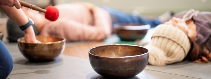 A person uses a red mallet to play a brass singing bowl. Several singing bowls sit on yoga mats with people lying relaxed in the background.