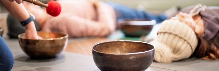 A person uses a red mallet to play a brass singing bowl. Several singing bowls sit on yoga mats with people lying relaxed in the background.