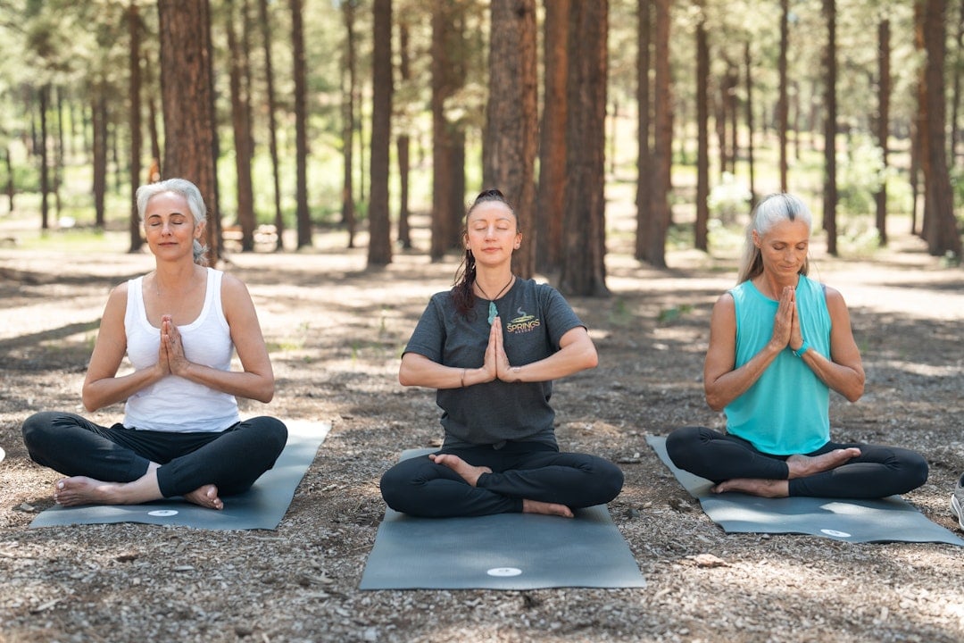 Three women meditate, eyes closed, hands in prayer pose, cross-legged on yoga mats in a sunlit pine forest. The middle woman's shirt reads: SPRINGS RESORT.