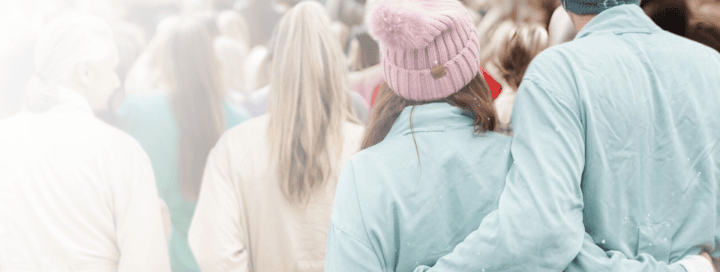 The backs of a couple, man's arm around woman's shoulder, walk amidst a blurred crowd. They wear winter hats as small white particles fall.