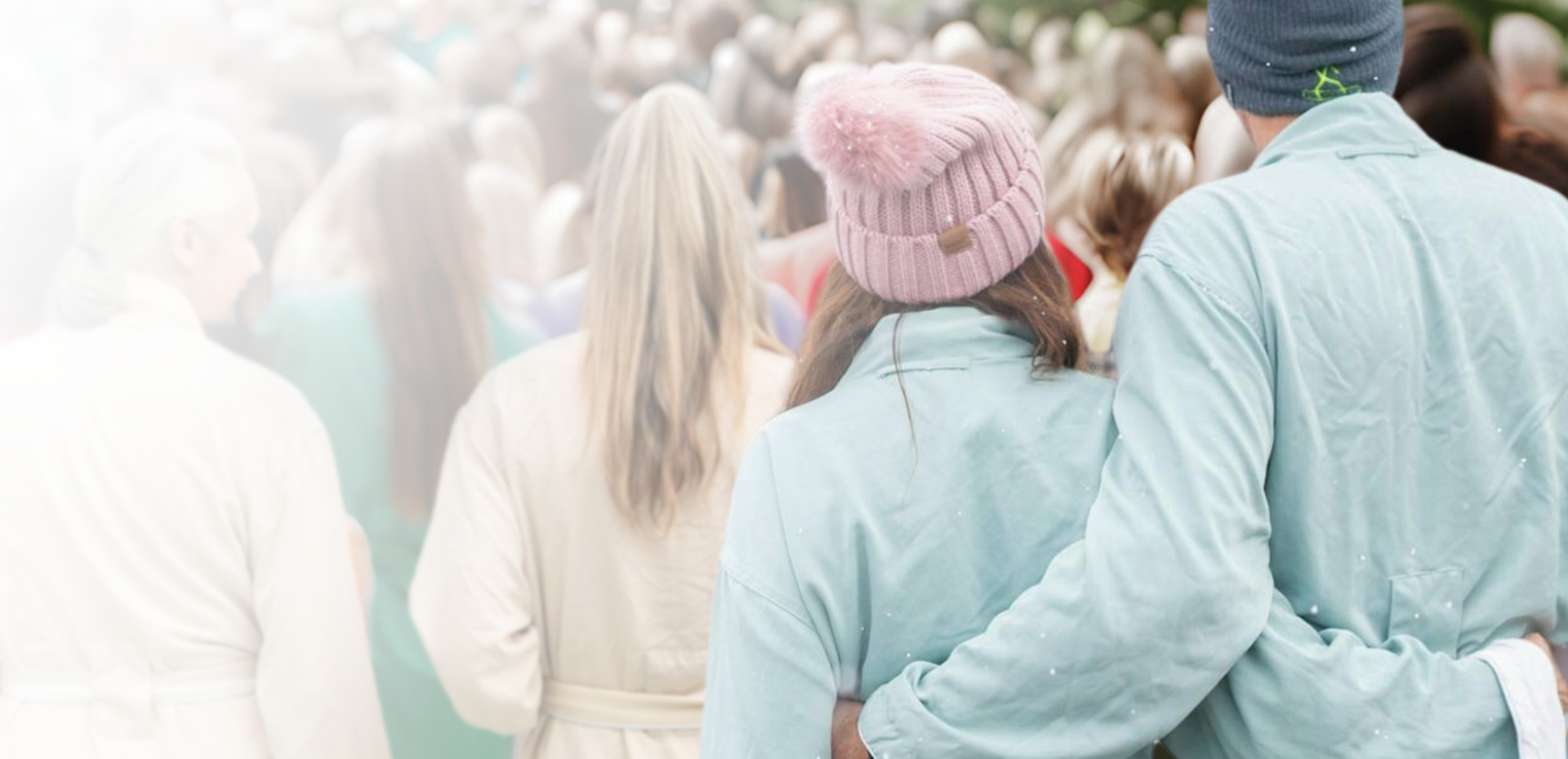 The backs of a couple, man's arm around woman's shoulder, walk amidst a blurred crowd. They wear winter hats as small white particles fall.