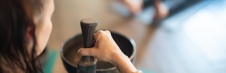 A woman's arm holds a wooden mallet over a bronze singing bowl in a blurred studio where another person lies.