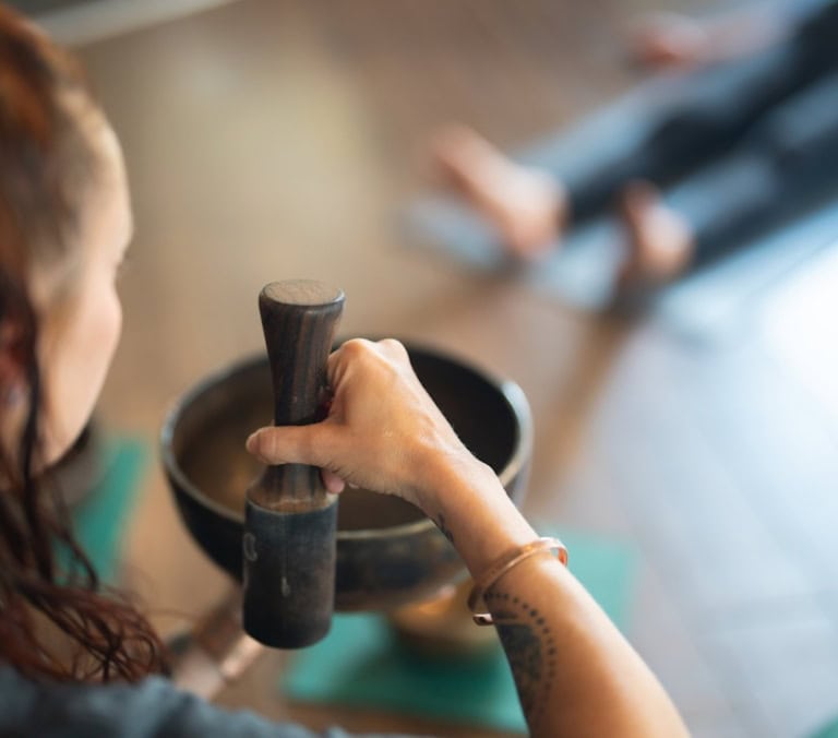 A woman's arm holds a wooden mallet over a bronze singing bowl in a blurred studio where another person lies.