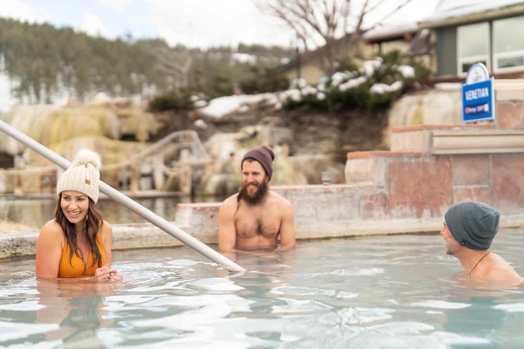 Three people smile while relaxing in an outdoor thermal pool, surrounded by snowy hills and buildings. A sign reads: VENETIAN