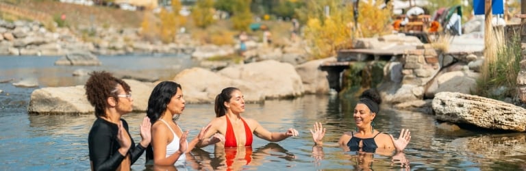 Four women smiling and gesturing with raised hands, standing chest-deep in an outdoor natural hot spring among rocks and autumn foliage.