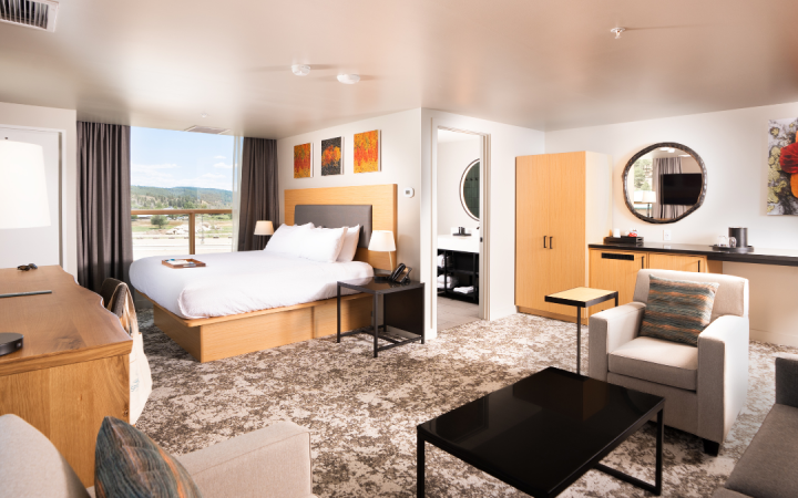 A spacious, modern hotel suite features a king bed, desk, and seating area. Large windows overlook a tree-lined valley, with a bathroom entry visible.