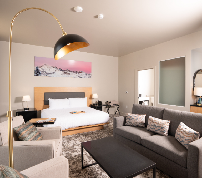A modern hotel suite shows a bed and a grey sofa facing a coffee table. A gold arched lamp illuminates an armchair on a patterned rug. A mountain artwork hangs above the bed.