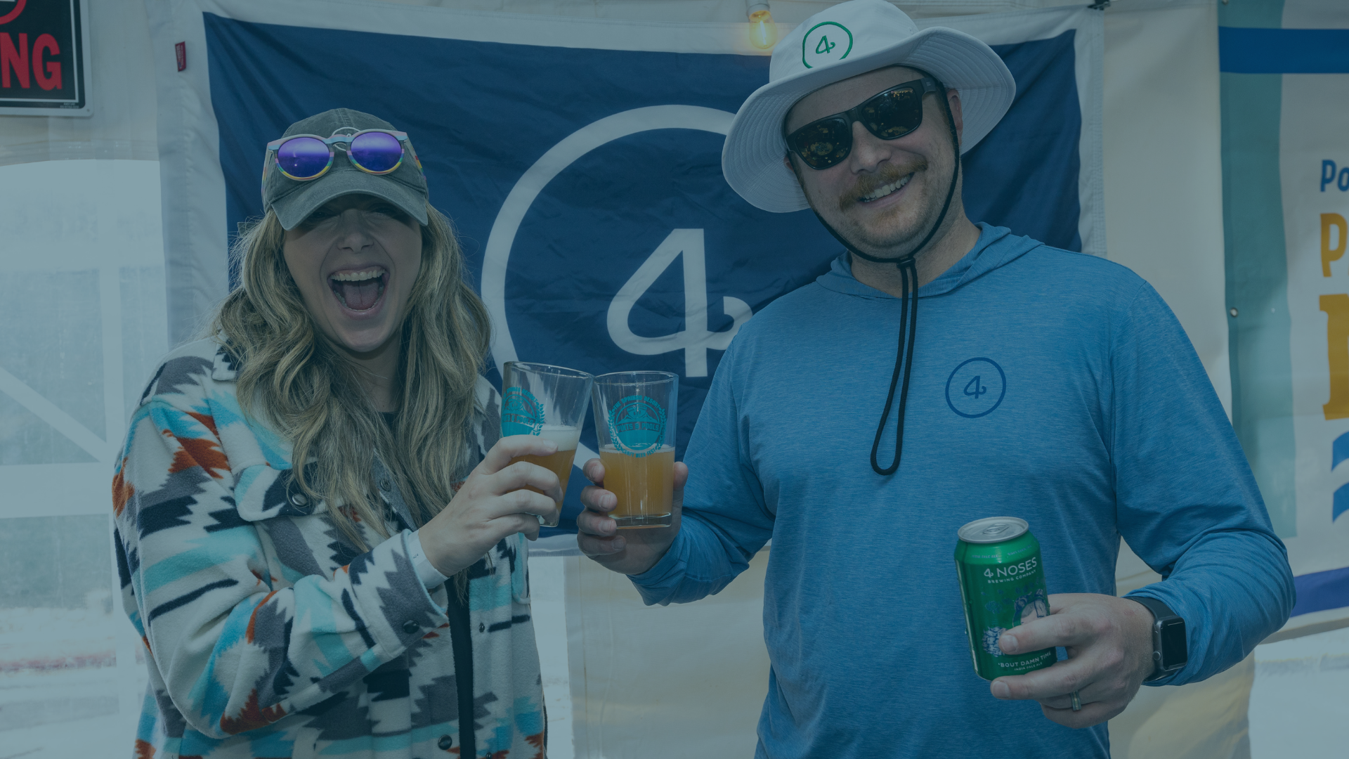 A smiling woman cheers with a beer glass, mouth wide open, while a man smiles, holding a beer glass and green can. They stand before a blue 