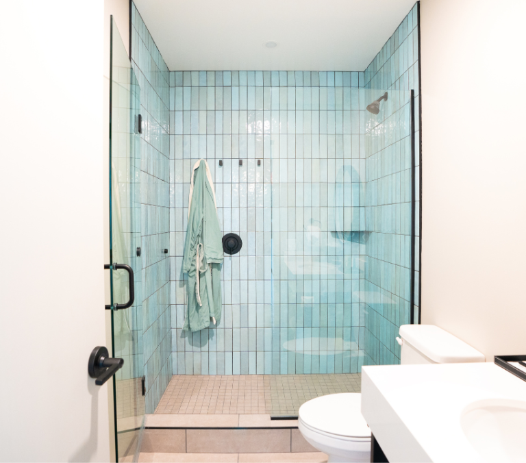 A light green robe hangs inside a glass shower with vertical light blue tiles. A white toilet and sink are visible to the right, creating a modern bathroom scene.