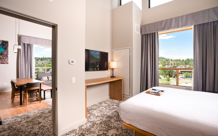 Modern hotel suite shows a bedroom with a bed, TV, and desk, opening to a living area with a dining table. Large windows reveal a scenic, hilly town. Text: 70.