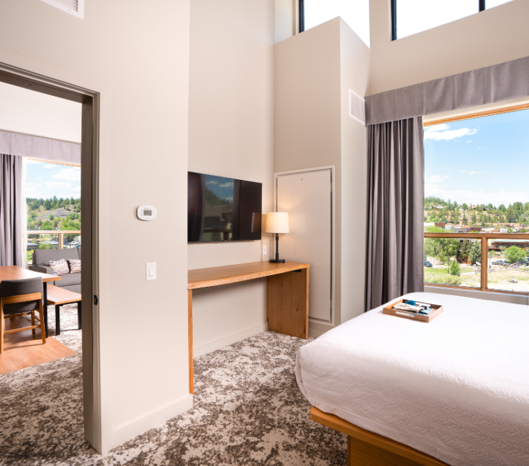 Modern hotel suite shows a bedroom with a bed, TV, and desk, opening to a living area with a dining table. Large windows reveal a scenic, hilly town. Text: 70.