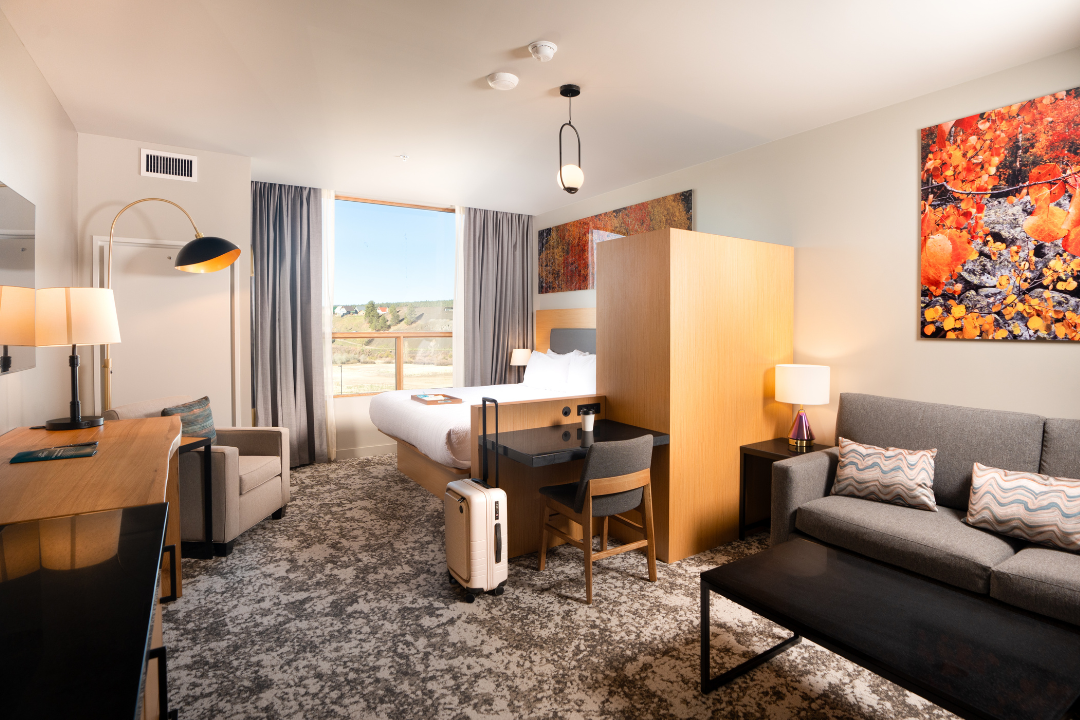 A modern hotel room features a bed with a tray, a desk, a sofa, and an armchair, set on a patterned carpet. A large window overlooks a natural landscape.
