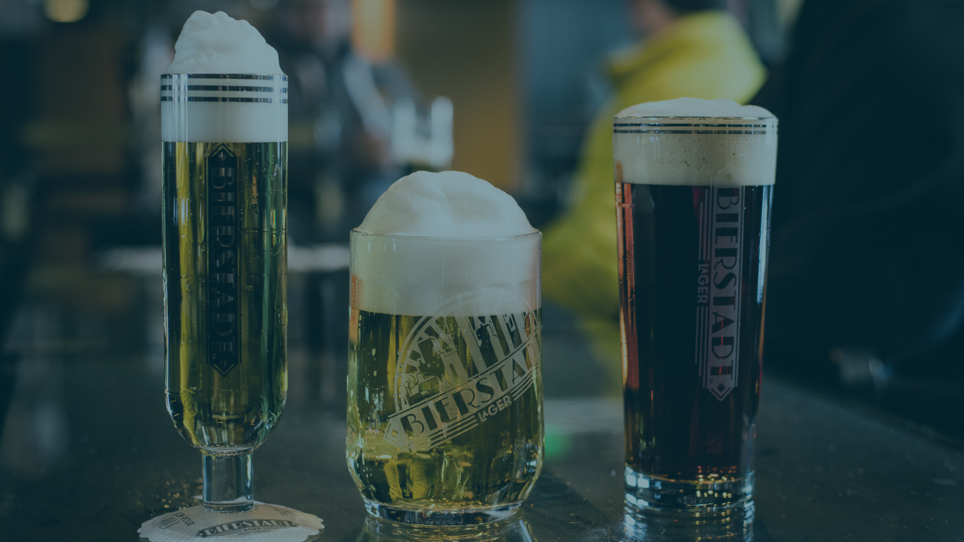 Three diverse glasses of beer, with white foam heads, are displayed on a dark, reflective bar counter, with blurry background patrons. Text: Left glass: BEERSTADER. Middle glass: BIERSTADT LAGER. Right glass: BIERSTADT LAGER. Coaster: BIERSTADT LAGER, MADE IN COLORADO.