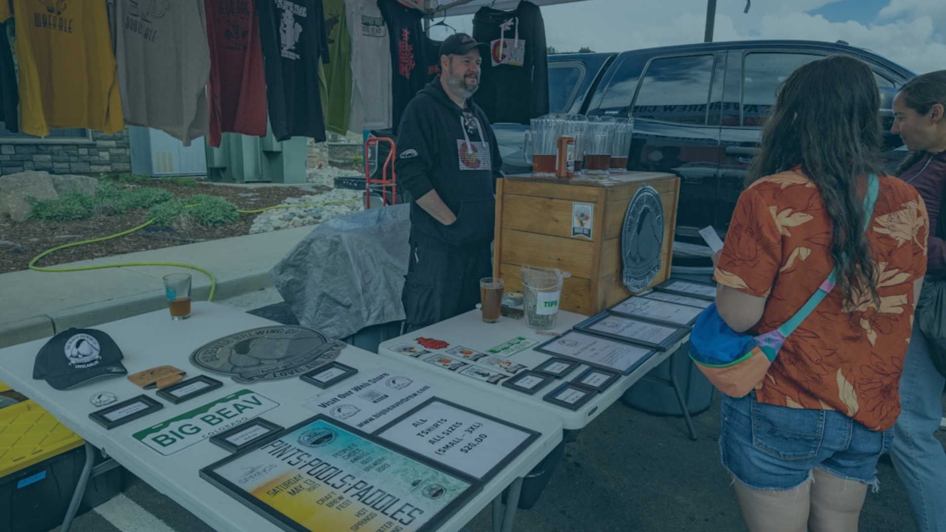 A male vendor stands behind a table displaying merchandise and beer, interacting with two female customers at an outdoor market.
