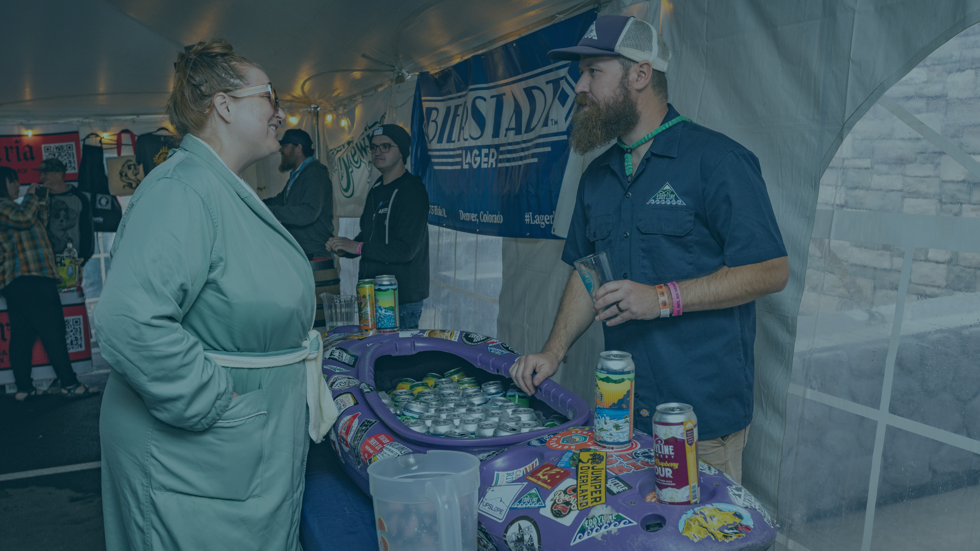 Woman in a green robe conversing with a bearded man in a blue shirt next to a purple canoe-shaped cooler filled with drinks, under a white tent. BIERSTADT™ LAGER, 5 bbl Denver, Colorado #Lager, EDDYLINE BREWERY MOUNTAIN TIME, JUNIPER OVERLAND, LINE PINE BREWERY Raspberry Sour.