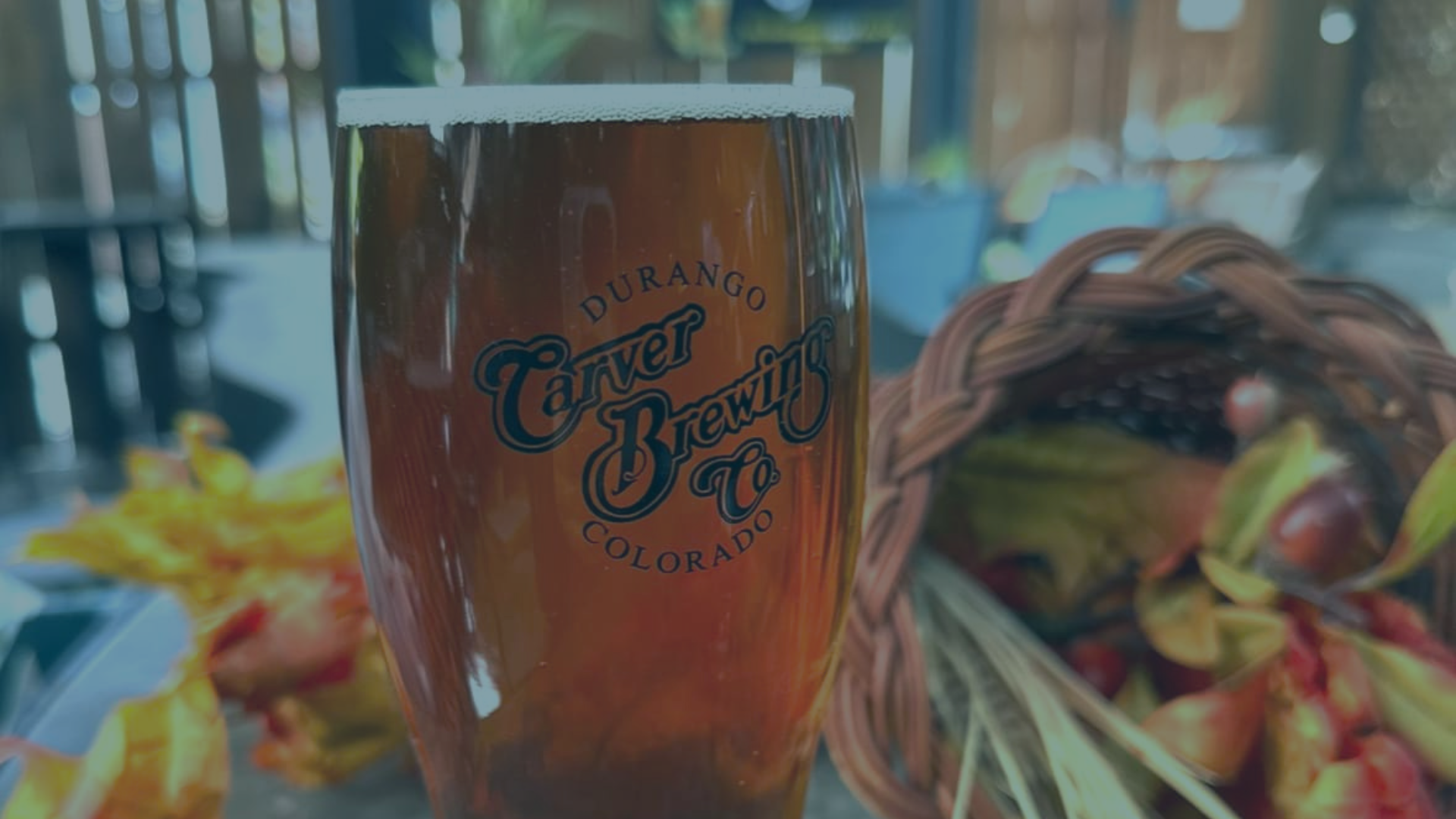 Amber beer in a branded pint glass on a table, beside a cornucopia of fall decor, on an outdoor patio. The glass reads: 