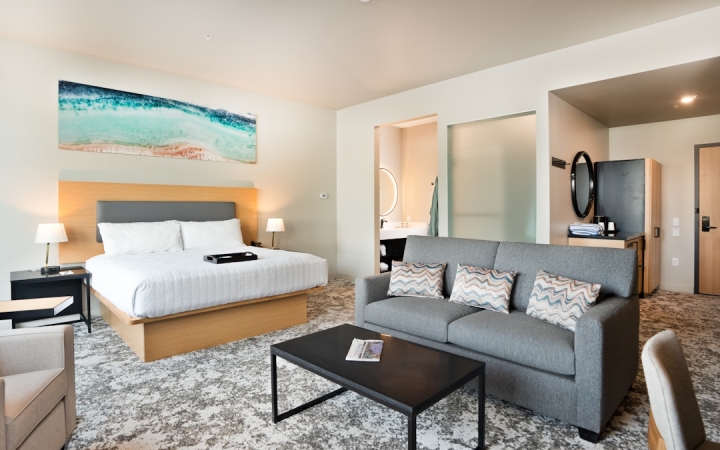 A modern hotel room features a king bed, grey sofa, and coffee table on patterned carpet. A bathroom with a round mirror is visible, alongside a wet bar near the room's entrance.