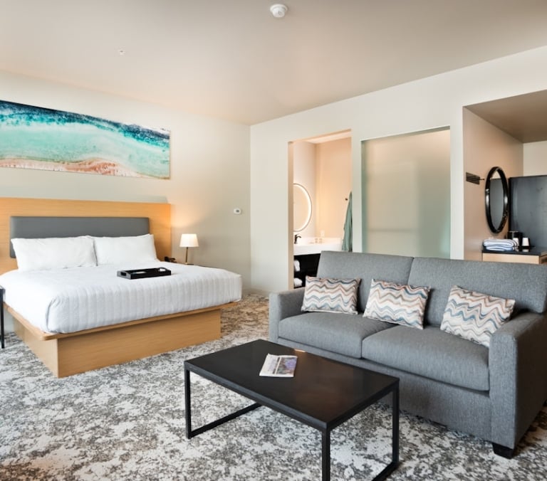 A modern hotel room features a king bed, grey sofa, and coffee table on patterned carpet. A bathroom with a round mirror is visible, alongside a wet bar near the room's entrance.